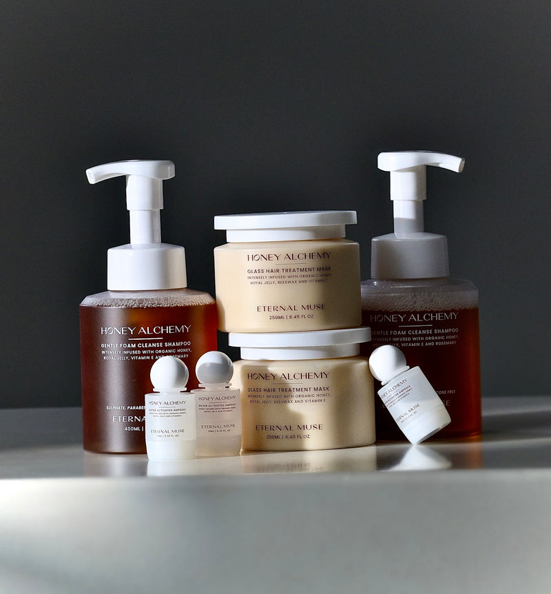 A set of hair care products including a shampoo, hair treatment mask, and gloss infusion ampoules, with pump dispenser bottles and a small glass jar with a white label, displayed on a reflective surface with a dark background.